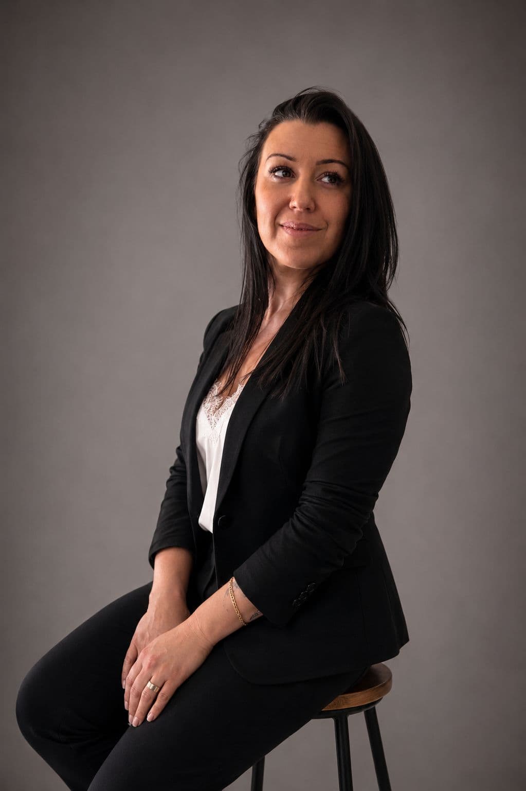 Jennifer Carles, assise sur un tabouret, en tailleur noir sur fond gris.