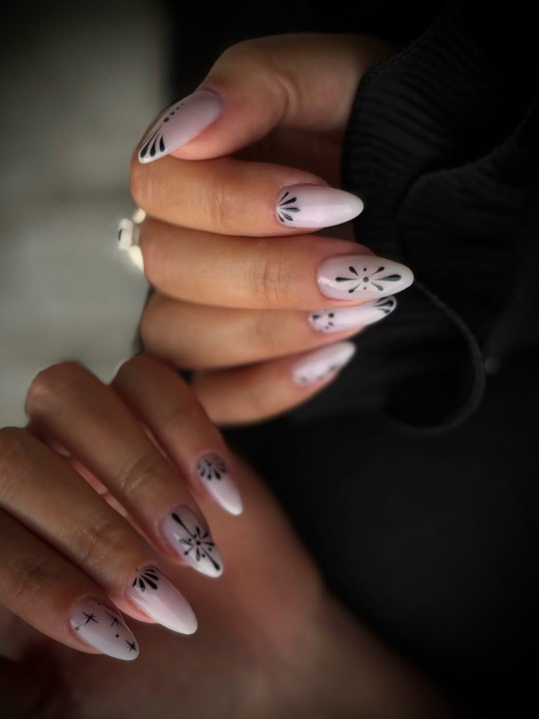 Deux mains aux ongles amande rose pâle, ornés de fins motifs noirs, sur fond sombre.